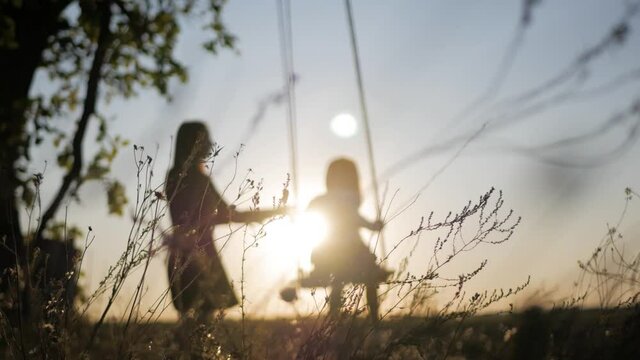 Happy young mother and little daughter on a swing at sun light. Pretty girl sitting on a wooden swing and looking at golden sunset. Happy family concept.