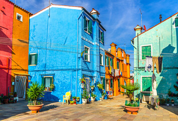 Fototapeta na wymiar Colorful houses of Burano island. Multicolored buildings and flowers in pots in small square, blue sky background in sunny summer day, Venice Province, Veneto Region, Northern Italy. Burano postcard