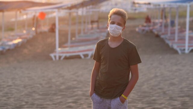 Portrait of young white kid wearing medical face mask while standing outdoor in summer beach of hotel resort. Boy looks into camera.