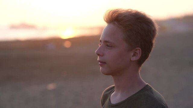 Closeup view 4k video portrait of cute handsome smiling happy young kid of 13 years old standing alone on scenic sunset sea beach.