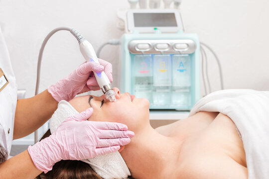 Beauty salon. The cosmetologist in medical gloves doing a hydra peeling procedure on the client's cheeks. Side view. Professional skin care and treatment concept