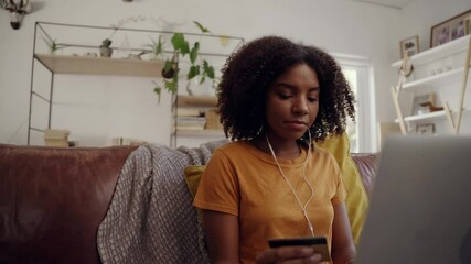 Smiling young woman sitting on cozy sofa with earphone in his ear holding credit card and typing on laptop making online payment - Powered by Adobe