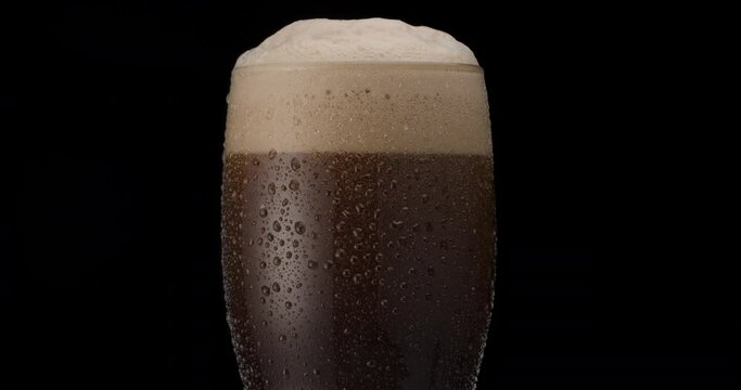 Glass of black beer on a black background. Beer sways in the glass, bubbles and foam rise. Glass of beer rotates slowly clockwise.