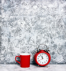 Red classic alarm clock and red coffee mug on gray plaster wall.