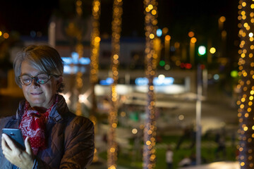 Smiling senior woman with glasses looking at her cellphone standing outdoor in the night with illuminated palm trees behind she. Christmas yellow lights decorations.