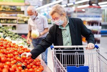 mature woman in mask and gloves with covid picks tomatoes in vegetable section of supermarket