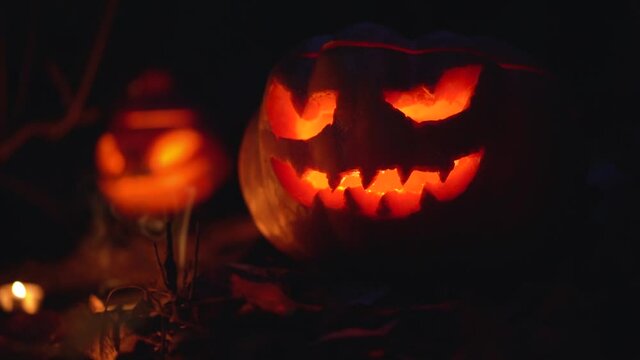 Two glowing creepy orange pumpkins with carved scary eyes, mouth in gloomy magic forest close up outdoors. Smoke from candles rises inside. Slow motion. Halloween horror concept. Copy space for text.