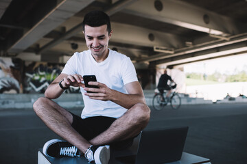 Fototapeta na wymiar Joyful man messaging on smartphone in skate park under bridge
