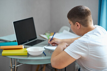 portrait of young caucasian teen boy studying at home, online studying, do homework during quarantine. sits in bedroom, wearing domestic clothes, alone