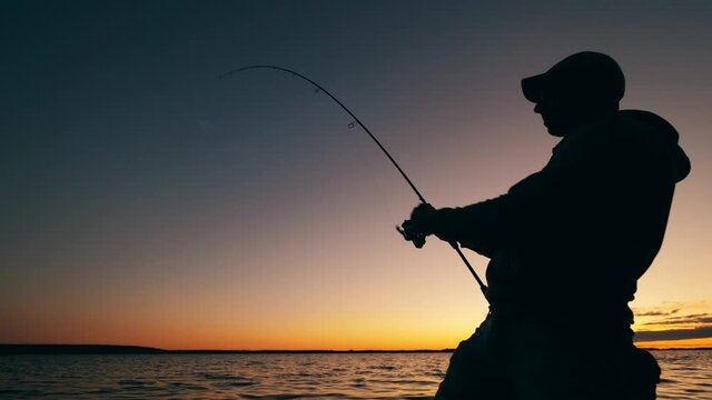 A man is reeling up a fishing pole in a sunset lake