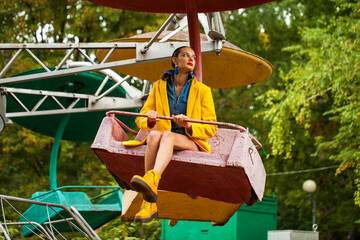 Fashion model in a bright yellow coat sits on a carousel in an amusement park