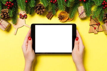 Top view of woman holding tablet in her hands on yellow background made of Christmas decorations. New Year holiday concept. Mockup
