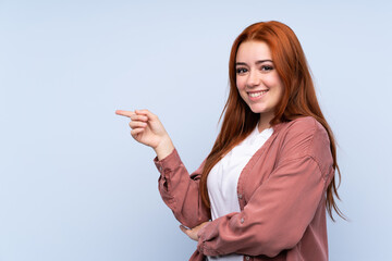 Redhead teenager girl over isolated blue background pointing finger to the side