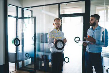 Confident male entrepreneurs discussing plans and finance strategy together talking in office, professional men looking at infographic on modern glass board while have productive communication