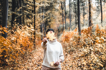 Woman covers her face with dry yellow leaf in autumn forest