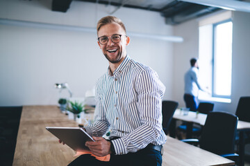 Portrait of cheerful male entrepreneur holding digital tablet and laughing at camera during work day in company office, satisfied businessman in optical eyeglasses rejoicing at table desktop