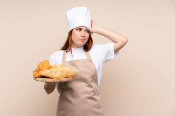 Redhead teenager girl in chef uniform. Female baker holding a table with several breads having doubts and with confuse face expression