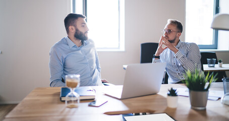 Fototapeta premium Skilled Caucasian colleagues in smart casual clothing collaborating at table desk with modern laptop technology for digital marketing, male employees discussing business information in office