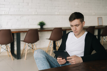 Handsome young man wearing casual fashion clothes sits at table and uses tablet. Relaxed freelancer working from home office sitting at a table with digital tablet.