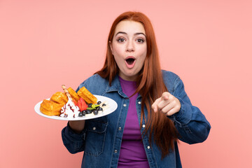 Redhead teenager girl holding waffles over isolated pink background surprised and pointing front