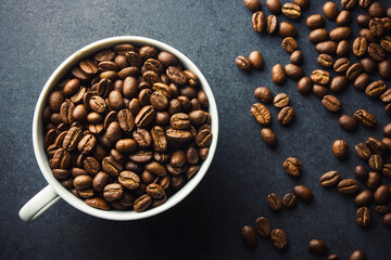 Coffee mug top view. White ceramic cup filled with coffee beans. Black table surface with coffee seeds. Morning caffeine background.