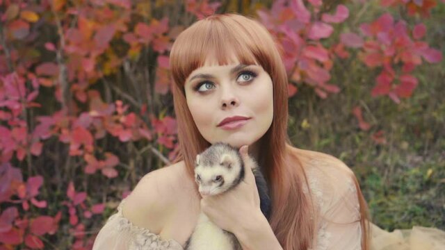 Portrait of a young beautiful woman who holds a little ferret in her arms. She looks with love and affection for her pet. Vintage medieval clothing on a background of autumn nature