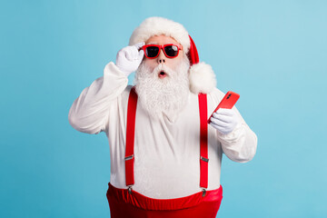 Fototapeta na wymiar Photo of pensioner grandfather grey beard hold telephone read fake news christmas cancelled wear santa x-mas red costume suspenders sunglass white gloves cap isolated blue color background