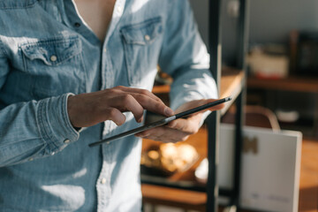 Close-up of hands of unrecognizable man using digital tablet pc while standing near shelves in office room, bright sunlight .