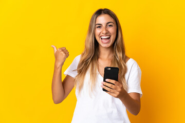 Fototapeta premium Young hispanic woman over isolated yellow background using mobile phone and pointing to the lateral