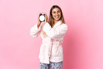 Fototapeta premium Young hispanic woman over isolated pink background in pajamas and holding clock with happy expression