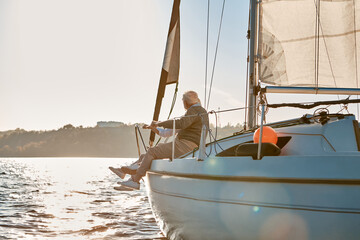 Enjoying sunset. Side view of a senior couple sitting on the side of a sailboat or yacht deck floating in a calm blue sea and looking at the horizon
