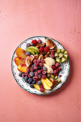 Assorted sliced fruits on vintage plate. Top view of mango, kiwi fruits, figs, grape, bananas, raspberries and blueberries on pink background.