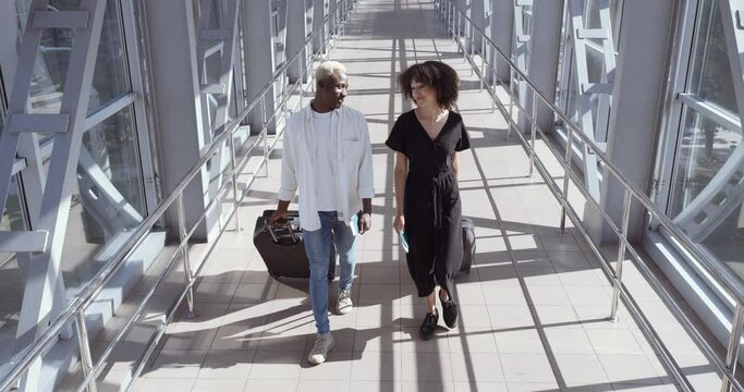 African woman and American black man walk together talking, carry suitcases, enter airport terminal or train station, meet two passengers before boarding, travel vacation together friends or marrieds