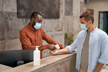Young african man wearing medical mask standing at reception in the modern office and measuring...