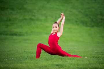 Obraz na płótnie Canvas schoolgirl girl in a bright red overalls is engaged in gymnastics on the grass in park, doing exercises for stretching