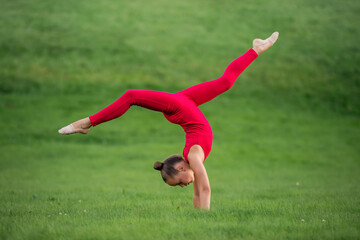 schoolgirl girl in a bright red overalls is engaged in gymnastics on the grass in park, doing exercises for stretching