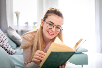 Happy woman with glasses reads book lying on comfortable sofa spending time at home