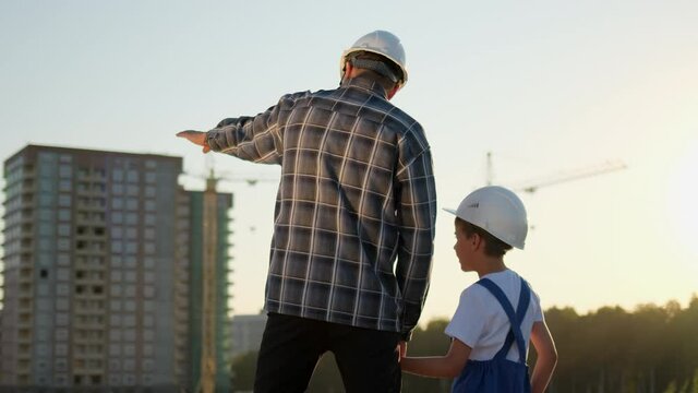 Father and son happy Family concept. Little boy and dad engineer builder architect with safety helmets, standing on background new buildings and construction cranes site. Future profession, 4 K slowmo