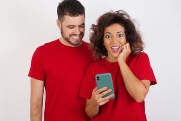 Joyous  Young beautiful couple wearing red t-shirt on wh poses with mobile phone device, types text message on modern smartphone, watches funny video during free time, enjoys good internet connection,