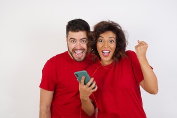 Positive Young beautiful couple wearing red t-shirt on white background holds modern cell phone connected to headphones, clenches fist from good emotions, exclaims with joy,