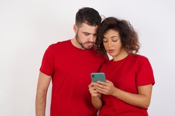 Portrait of excited Young beautiful couple wearing red t-shirt on white background  winking and eye hold smart phone use read social network news