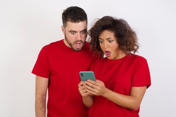 Portrait of focused Young beautiful couple wearing red t-shirt on white background use smartphone reading social media news, or important e-mail