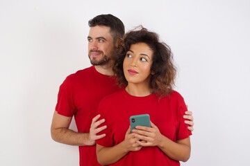 Photo of  Young beautiful couple wearing red t-shirt on white background holds telephone hands reads good youth news look empty space advert