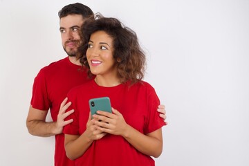 Young beautiful couple wearing red t-shirt on white background hold telephone hands read good youth news look empty space advert