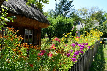 View of a small wooden hut or shack with an angled thatch roof located behind a beautifully maintained flower and herb garden with a dense orchard behind the object seen in Poland in summer