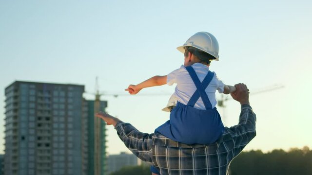 Father and son happy Family concept. Little boy and his dad engineer builder architect riding back with safety helmets, new buildings and construction cranes on site. Future profession, 4 K slow-mo