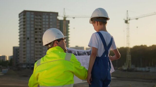 Father and son happy Family concept. Little boy and his dad engineer builder architect with safety helmets, showing new buildings and construction cranes on site. Future profession dream, 4 K slow-mo