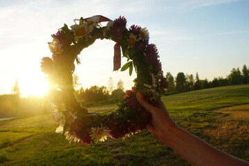 Female hands holding a wreath of wild flowers and a red and white ribbon with signs on a background of evening sunset