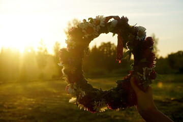 Female hands holding a wreath of wild flowers and a red and white ribbon with signs on a background of evening sunset