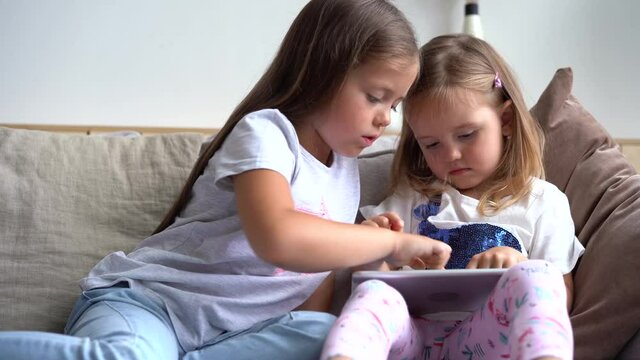 Cute little sisters sitting close to each other, watching something on the tablet device or playing a game on it.